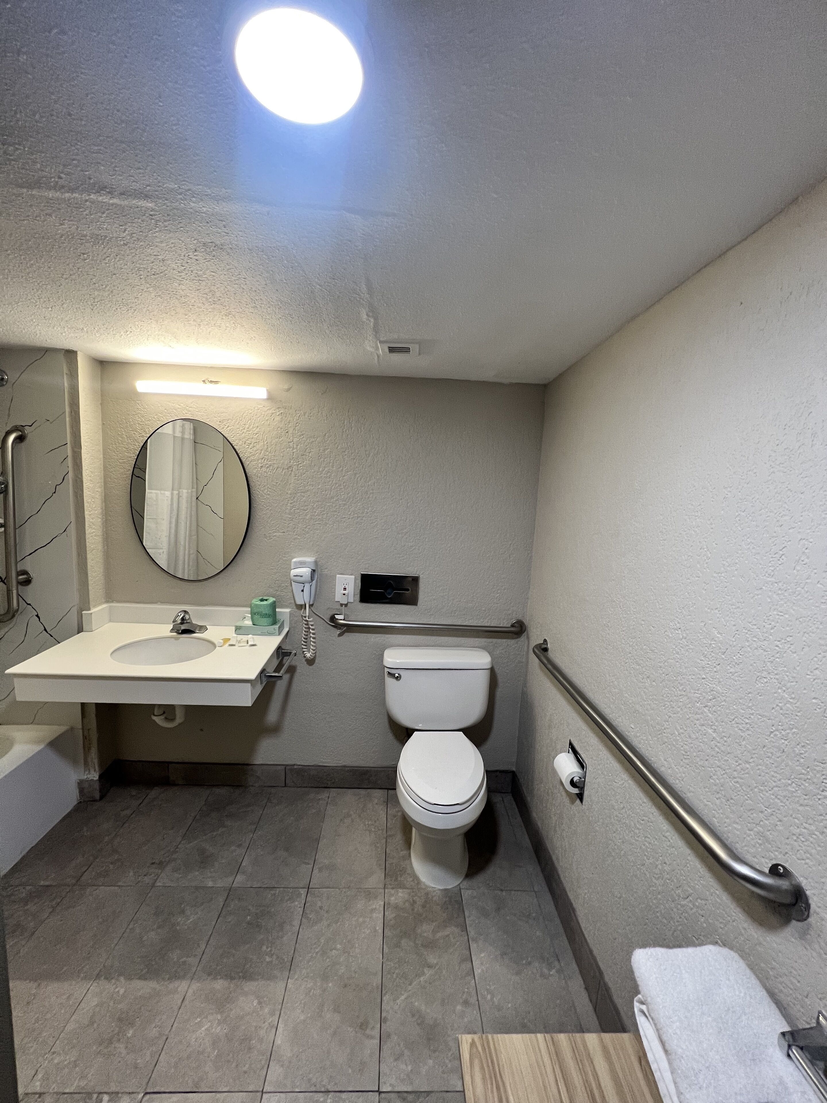 Un baño pequeño y funcional con lavamanos, inodoro y espejo. Incluye un secador de manos y un perchero para toallas.