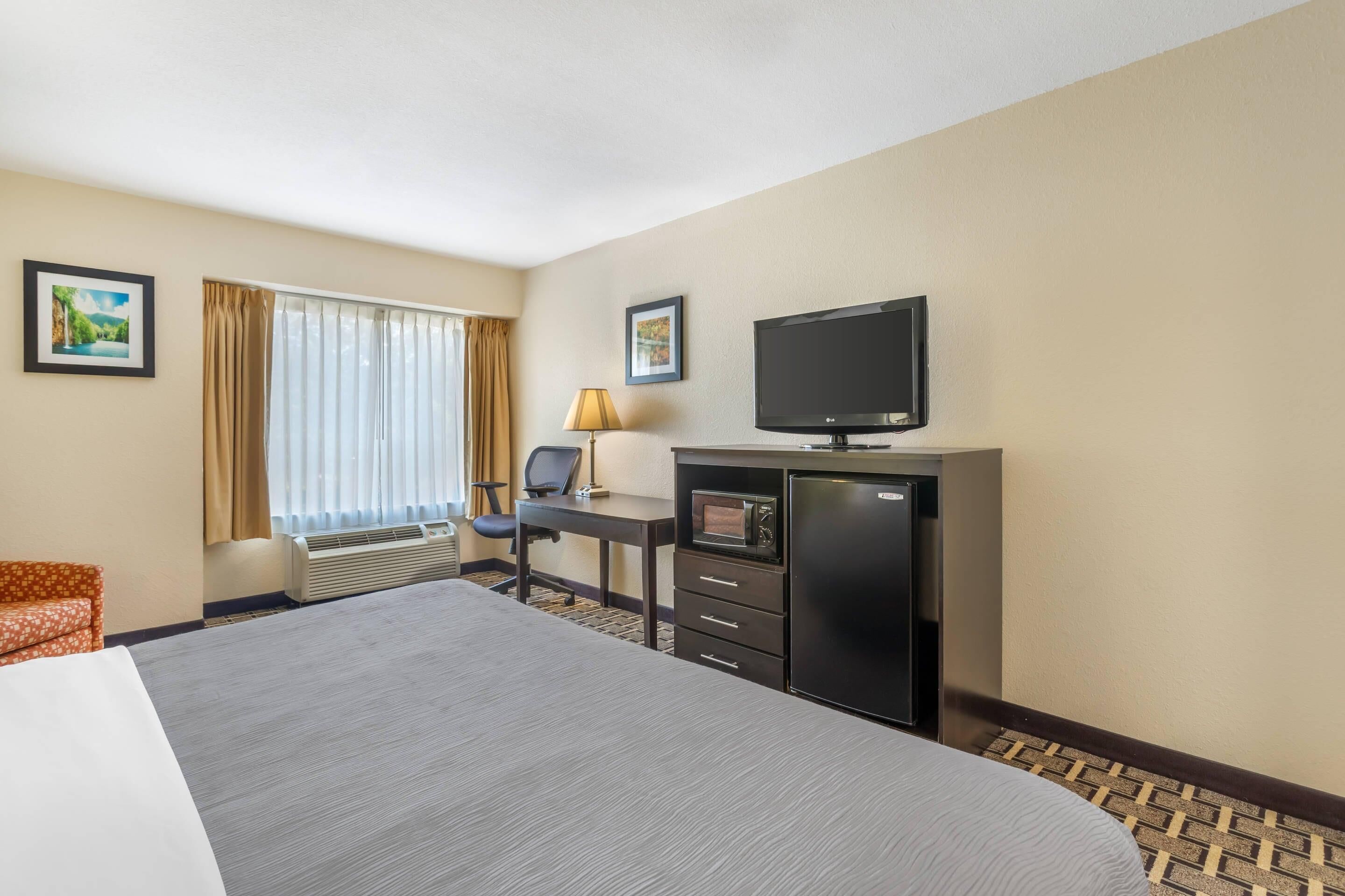 A hotel room with a large bed, a desk with a chair, a television, a mini-fridge, and a window with curtains.