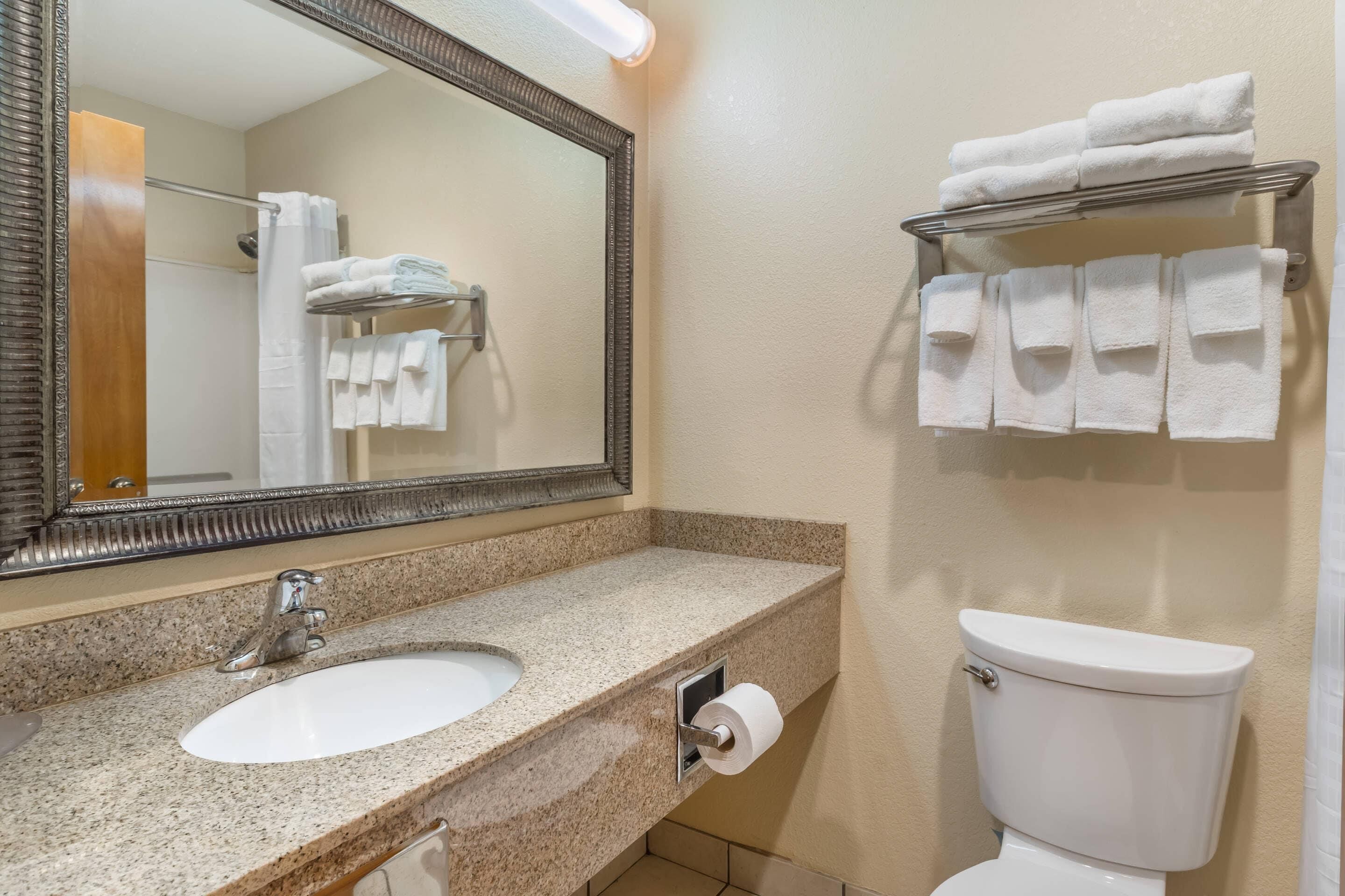 A bathroom with a granite countertop sink, a mirror, a toilet, and a towel rack with folded towels.