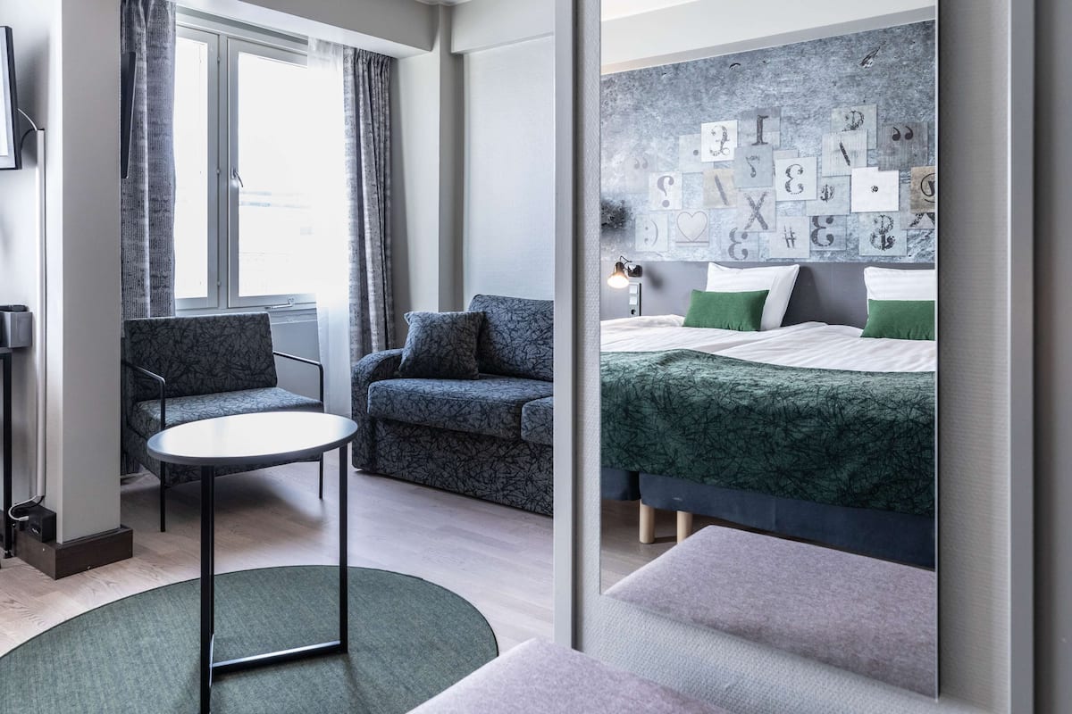 A modern hotel room with a bed, two sofas, a small table, and a view of the city through large windows.