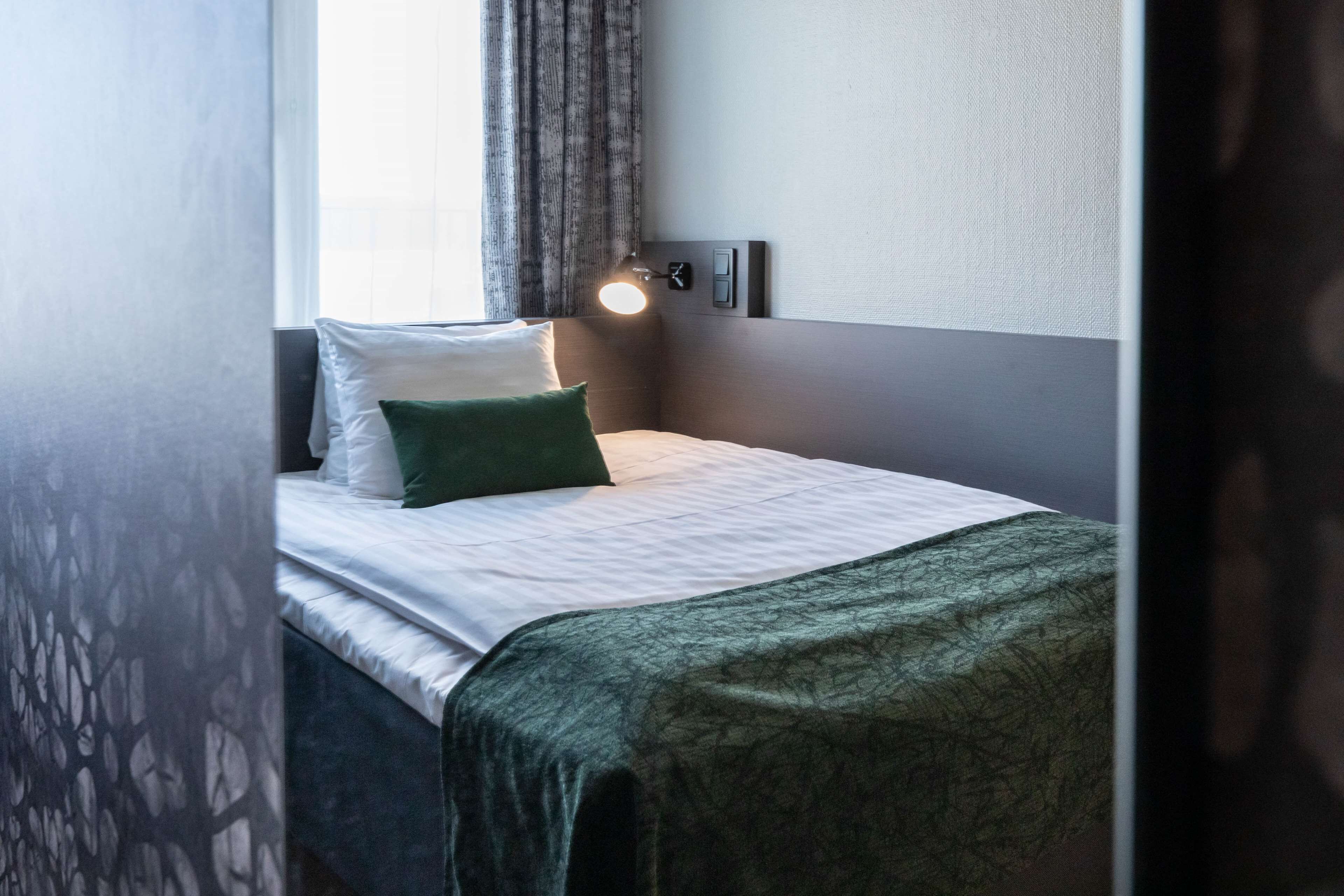 A neatly made bed with a green pillow and a white pillow, a bedside lamp, and a window with curtains.
