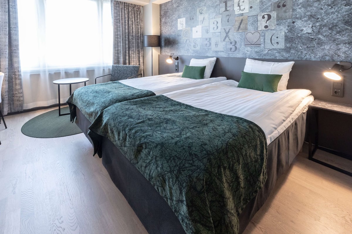 A hotel room with two beds, a small table, a lamp, and a wall with various symbols.