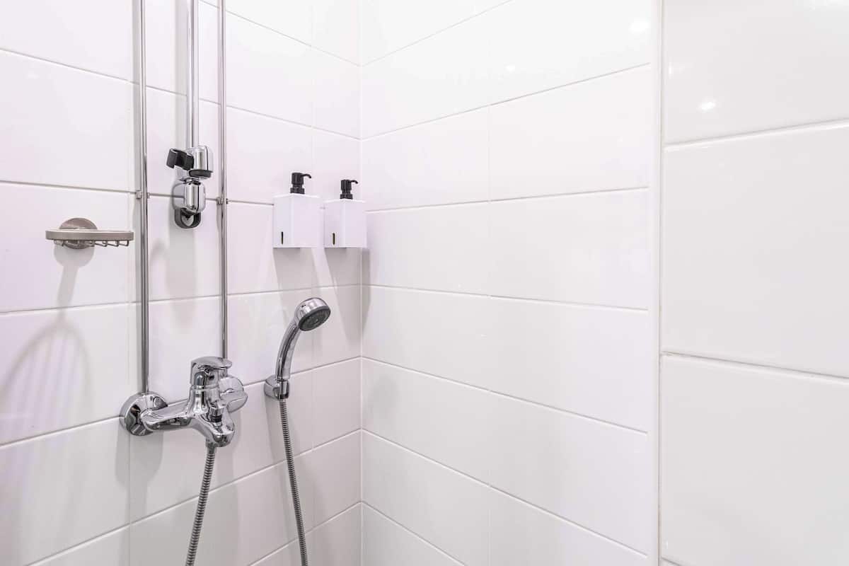 A bathroom with white tiled walls, a shower with a handheld showerhead, and two wall-mounted soap dispensers.