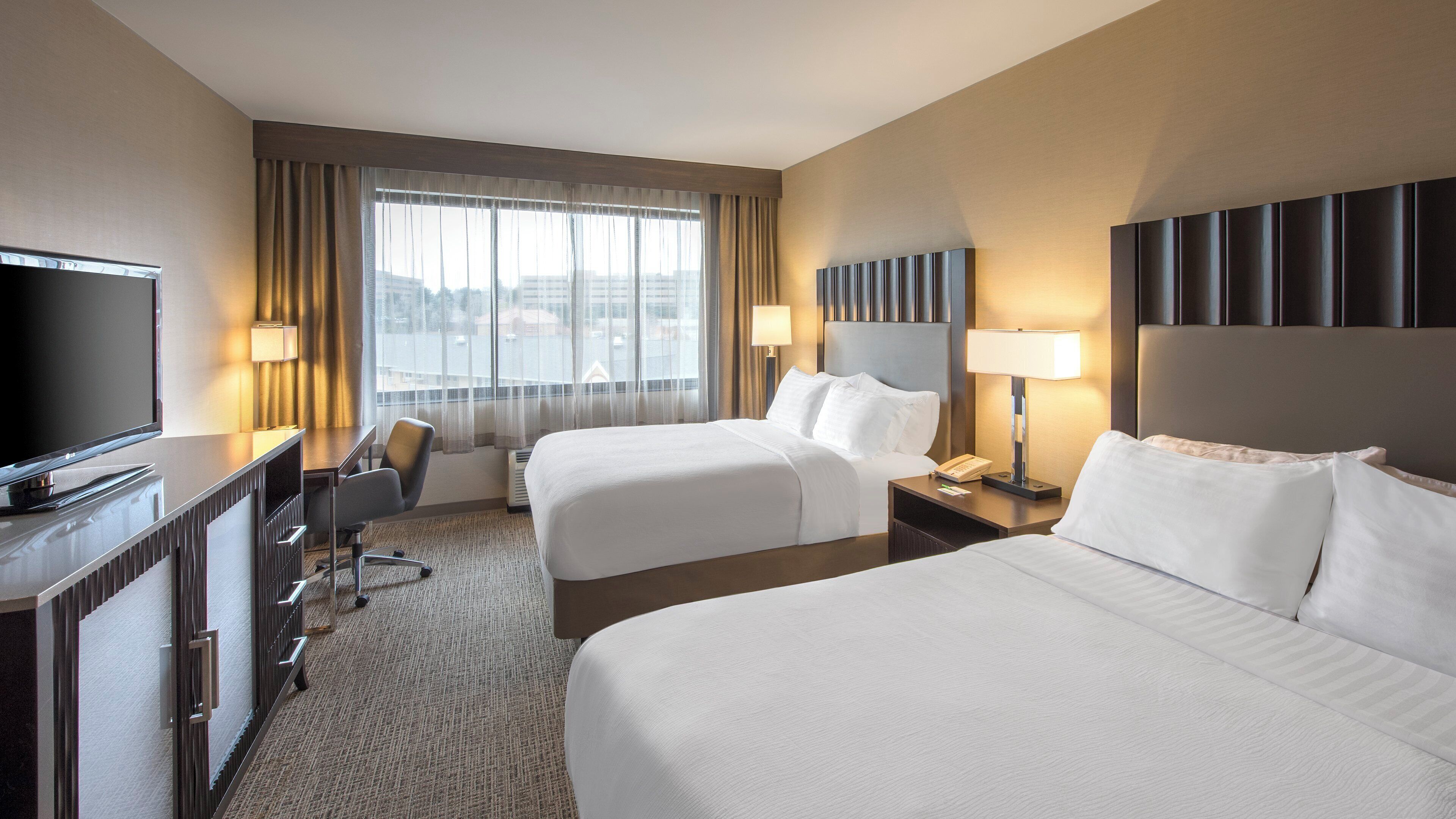Quarto de hotel com duas camas, uma escrivaninha com televisão, uma cadeira, um abajur e vista para a cidade.