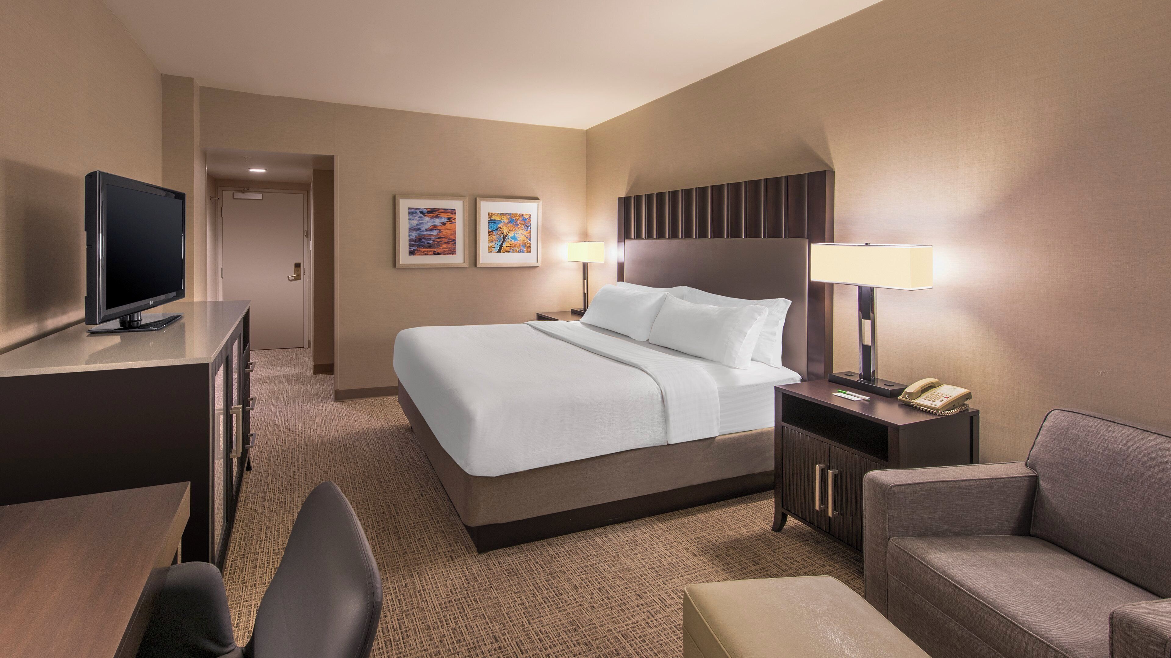 A hotel room with a bed, a TV, a desk, a chair, a couch, and a nightstand with a lamp and phone.