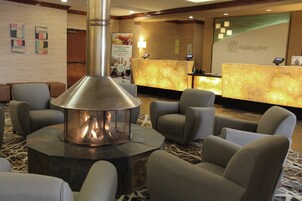 A lobby area with a central fire feature surrounded by seating, a reception desk, and a wall with a Holiday Inn sign.
