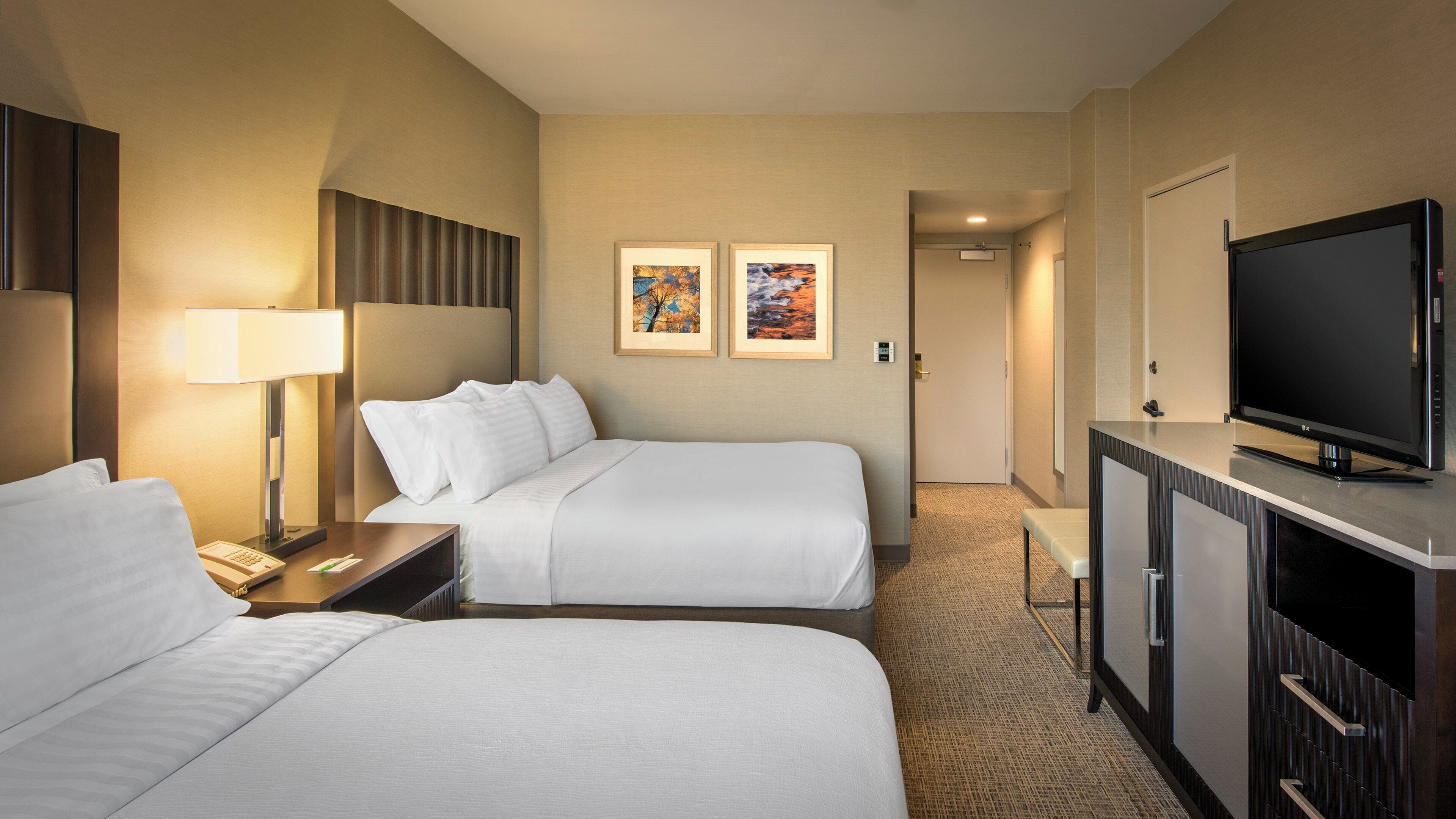 A hotel room with two beds, a desk with a lamp, a television, and two framed pictures on the wall.