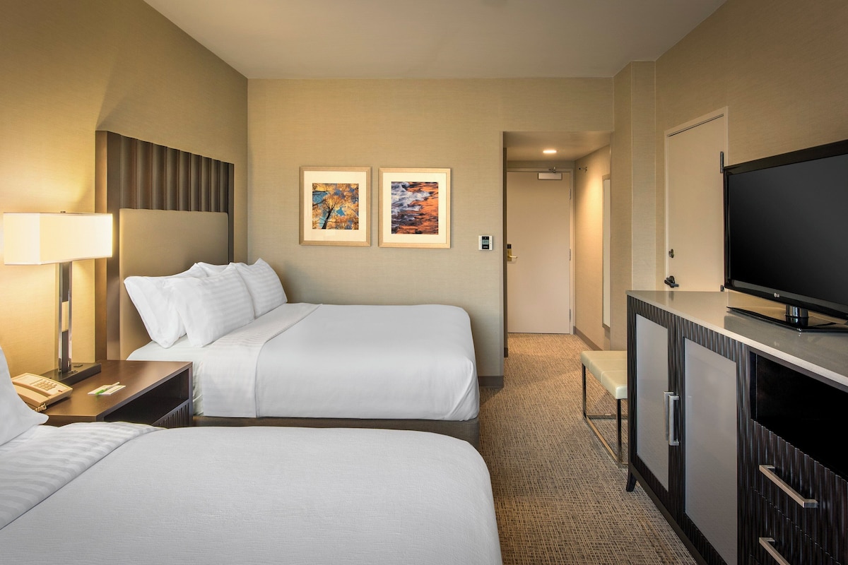 A hotel room with two beds, a desk with a lamp, a television, and two framed pictures on the wall.