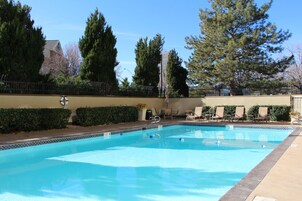 Una piscina con borde de azulejos, rodeada por una valla, junto a un edificio de pared color beige y un seto.