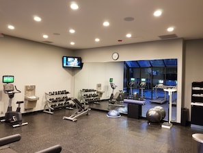 A gym with various exercise equipment, a television displaying workout videos, and a clock on the wall.
