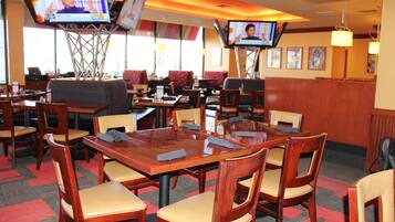 A restaurant with multiple tables and chairs, television screens mounted on the walls, and a patterned carpet.