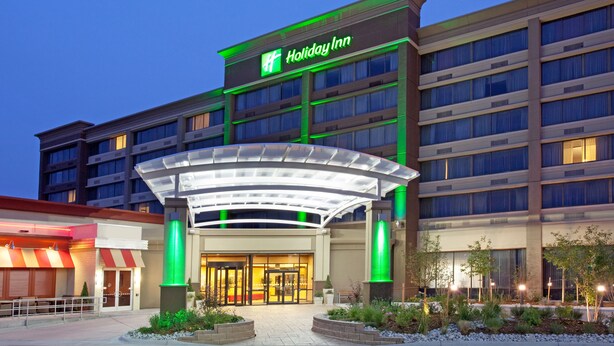Un hotel Holiday Inn dal design moderno, con un ingresso in evidenza caratterizzato da una tettoia in vetro e illuminazione verde.
