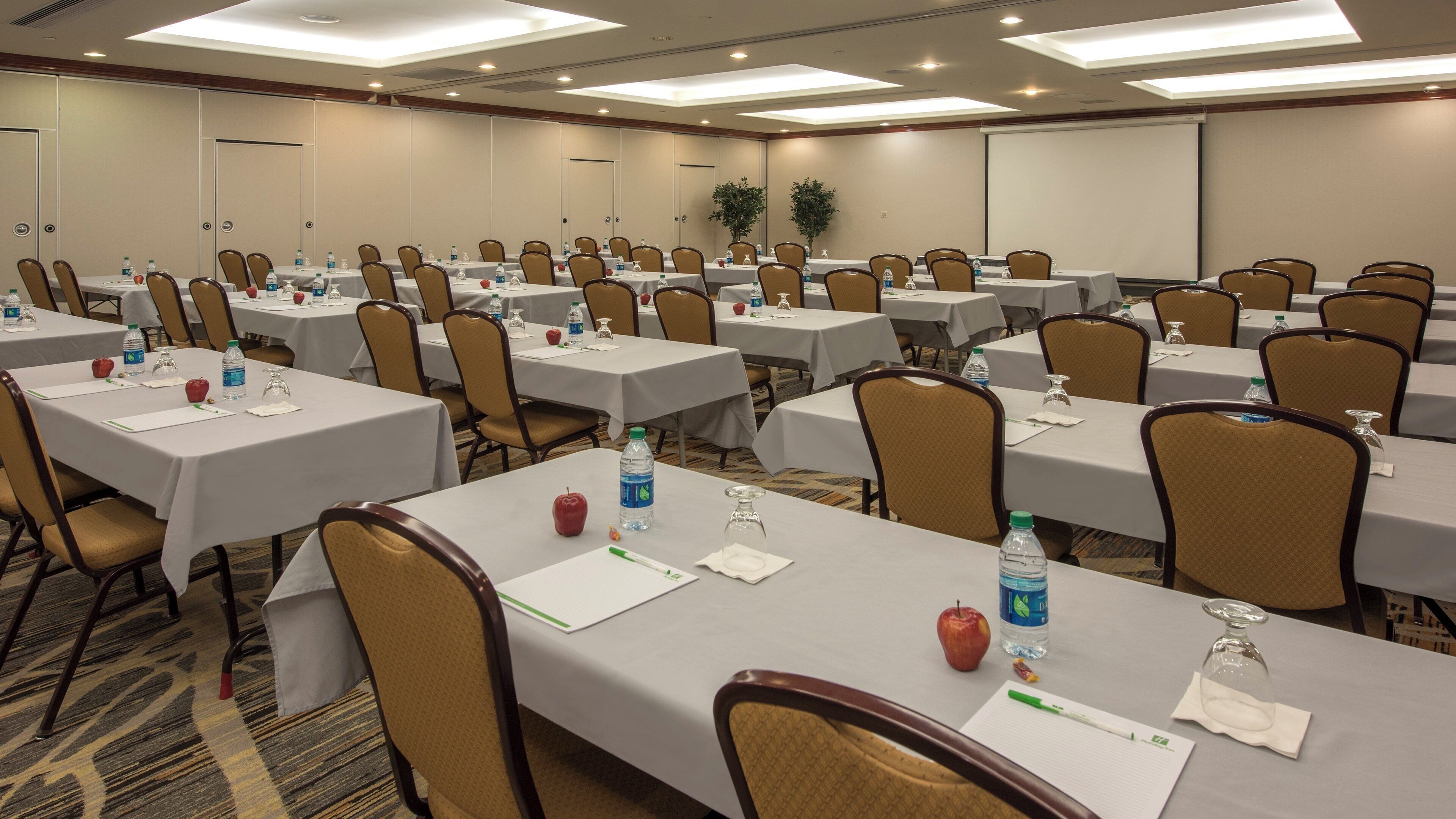 Un salón de conferencias con varias mesas, sillas, botellas de agua, manzanas y una pantalla de proyector.