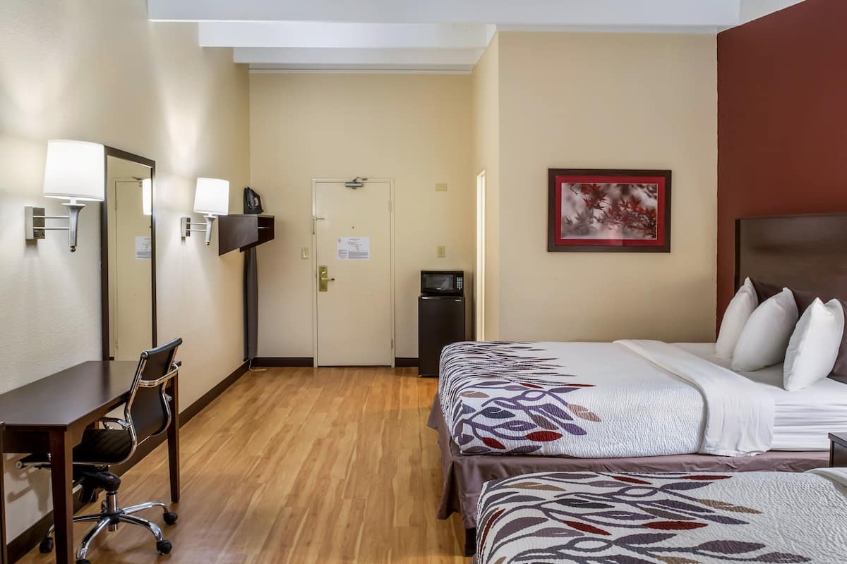 A hotel room with a bed, a desk with a chair, a small trash bin, and a door.