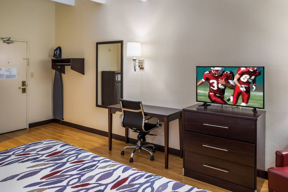 A hotel room with a bed, a desk with a chair, a TV, a dresser, a mirror, and a lamp.