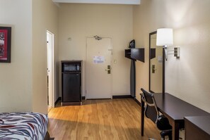 A hotel room with a bed, a desk, a chair, a refrigerator, a door, and a wall-mounted mirror. - Red Roof Inn Richmond, IN (Richmond)