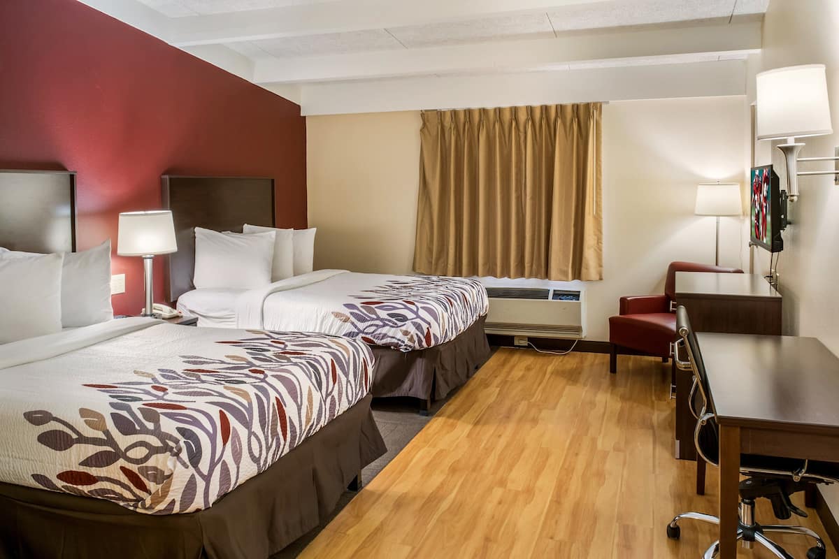 A hotel room with two beds, a desk, a chair, an air conditioning unit, and a television.