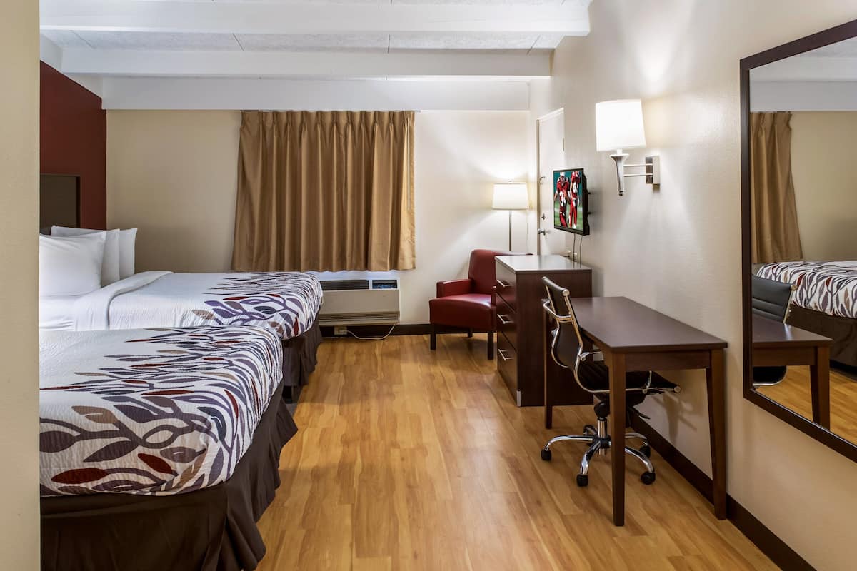 A hotel room with two beds, a desk, a chair, a desk lamp, a mirror, and a wall-mounted TV.