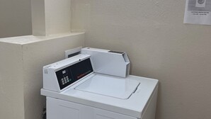 A laundry room with a front-loading washing machine and a wall-mounted dryer. There are two illuminated wall sconces above the machines. A sign indicating laundry room hours is visible on the wall. - Red Roof Inn Richmond, IN (Richmond)