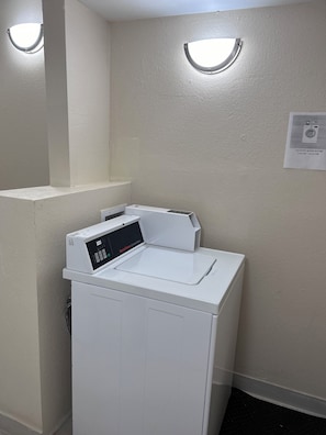 A laundry room with a front-loading washing machine and a wall-mounted dryer. There are two illuminated wall sconces above the machines. A sign indicating laundry room hours is visible on the wall. - Red Roof Inn Richmond, IN (Richmond)