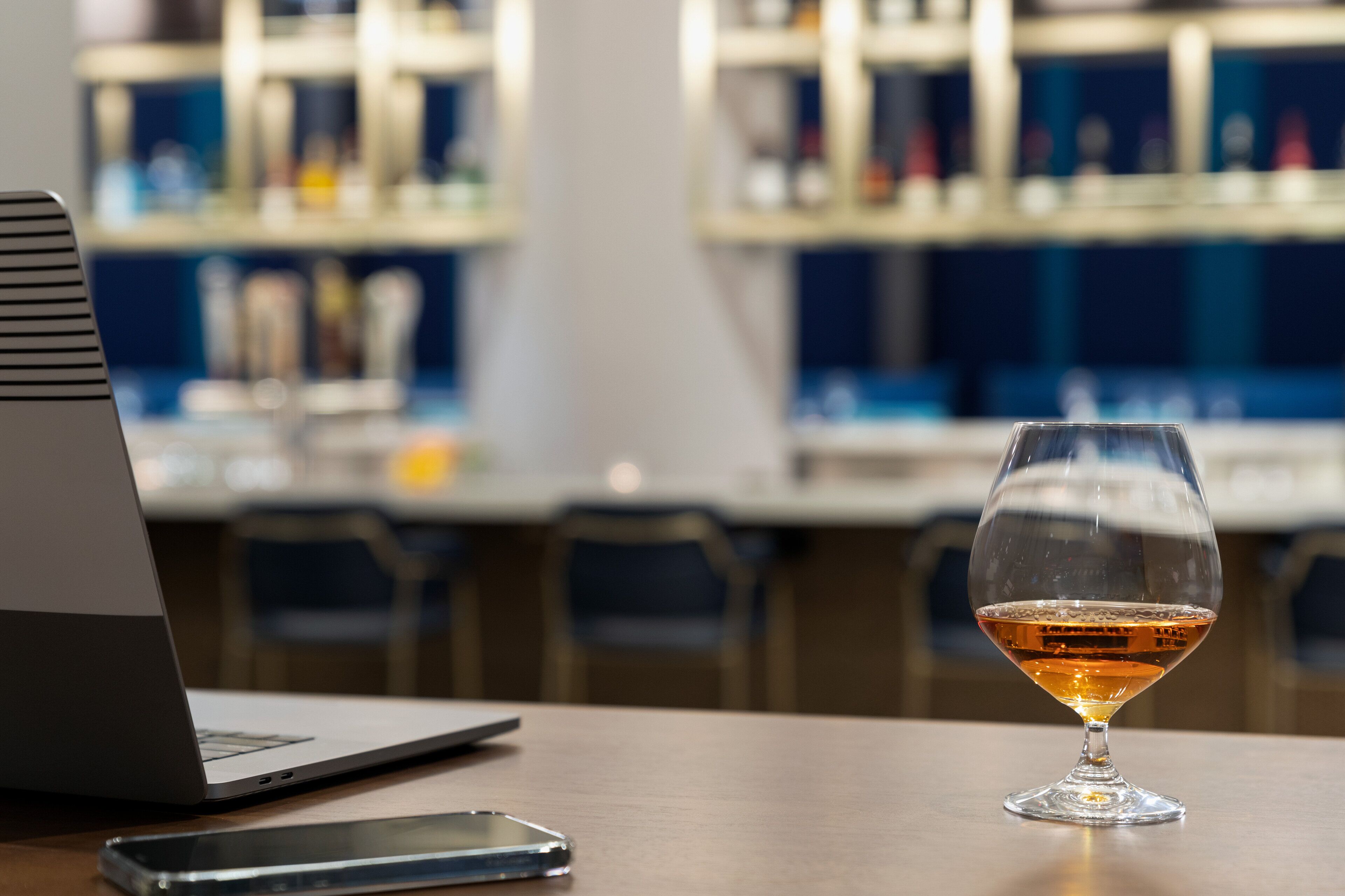 A laptop, a smartphone, and a glass of amber liquid on a wooden table in a bar setting.