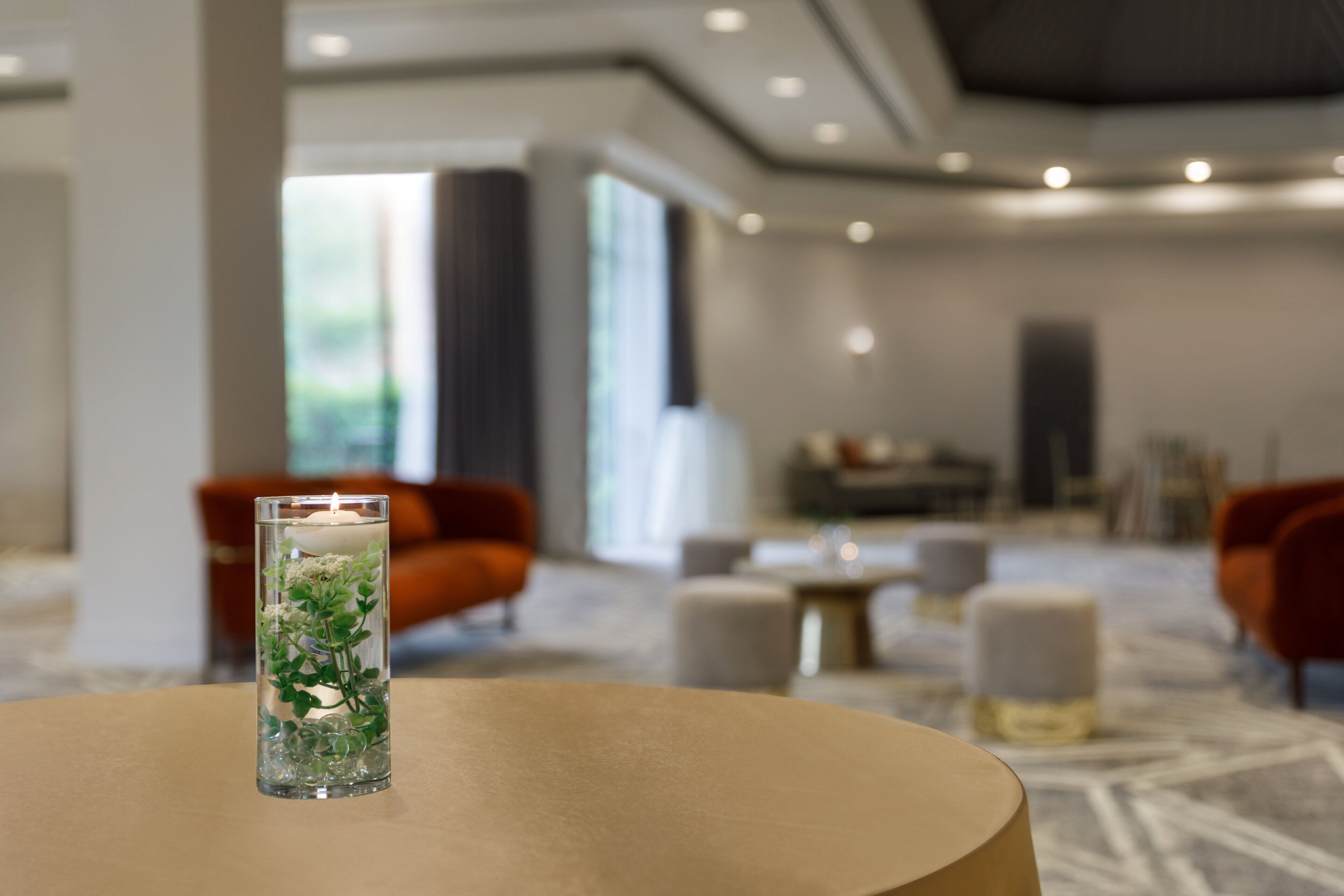 A modern hotel lobby with a round table in the foreground, a tall glass vase with greenery, and a seating area with orange chairs and a white ottoman in the background.