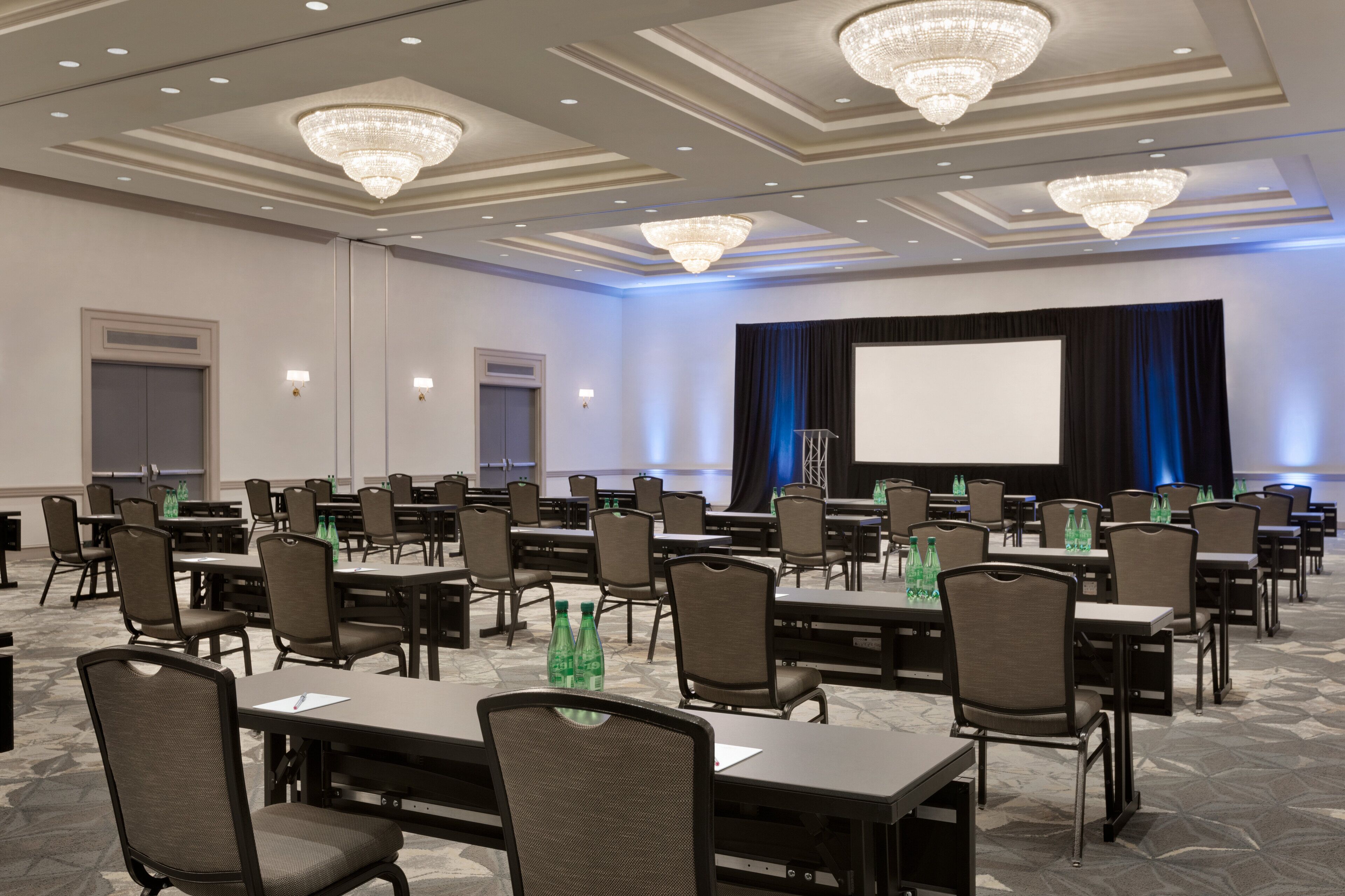 A conference room with rows of tables and chairs, a large screen, and chandeliers.