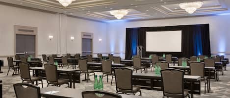 A conference room with rows of tables and chairs, a large screen, and chandeliers.