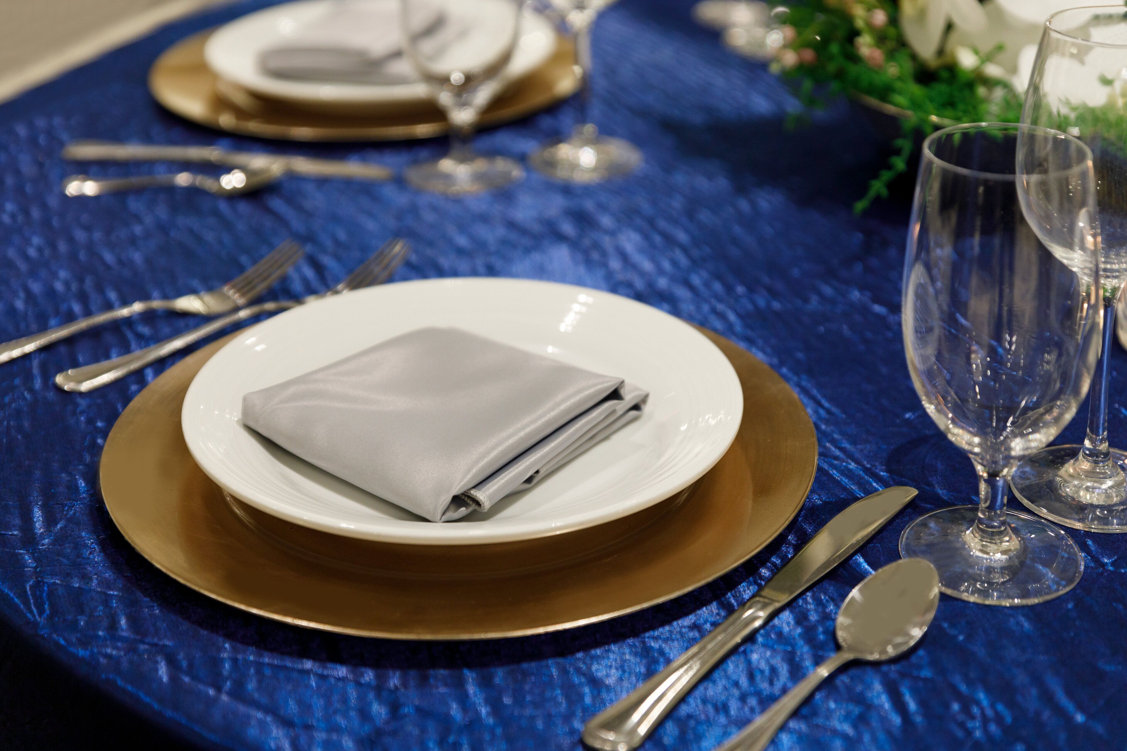 A table set with a folded napkin, a white plate, a gold charger, a knife, a spoon, and two wine glasses.