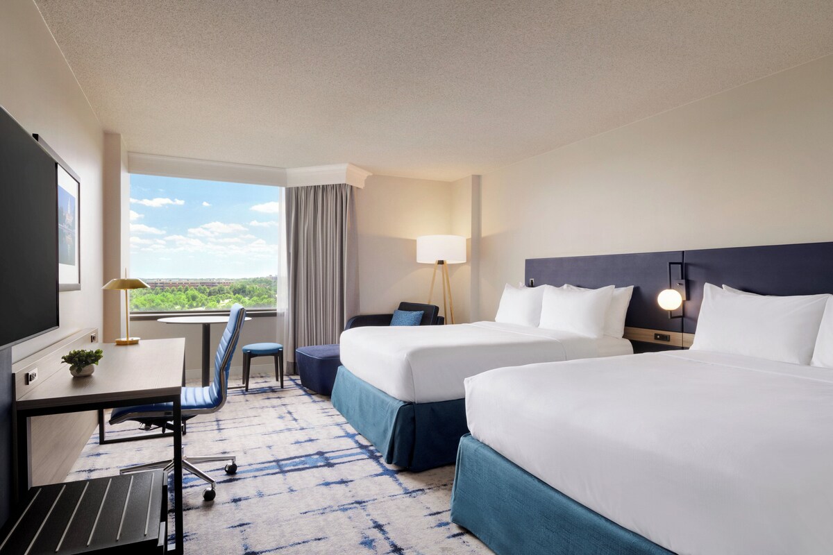 Een hotelkamer met twee bedden, een bureau met lamp, een stoel en uitzicht op het omliggende landschap.