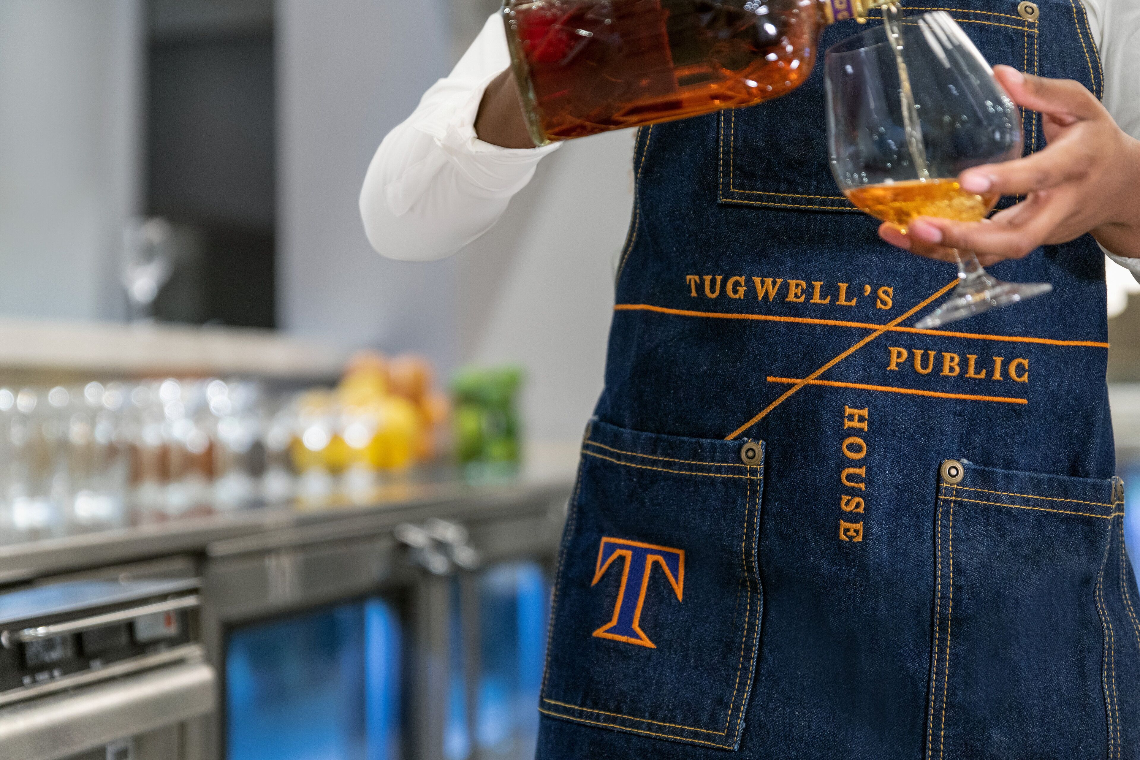 Une personne portant un tablier en jean sur lequel est imprimé le texte « TUGWELL’S PUBLIC HOUSE », tenant un verre et versant une boisson.