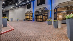 A modern hotel lobby with a brick floor, large planters with flowers, and a glass entrance.