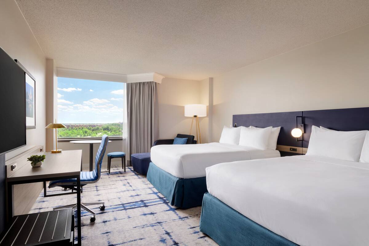 Een hotelkamer met twee bedden, een bureau met lamp, een stoel en uitzicht op het omliggende landschap.