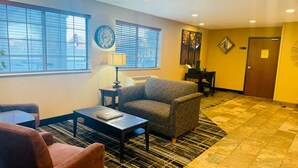 A hotel lobby with a sofa, armchair, and a small table with a book. There are windows with blinds, a clock, and a door.