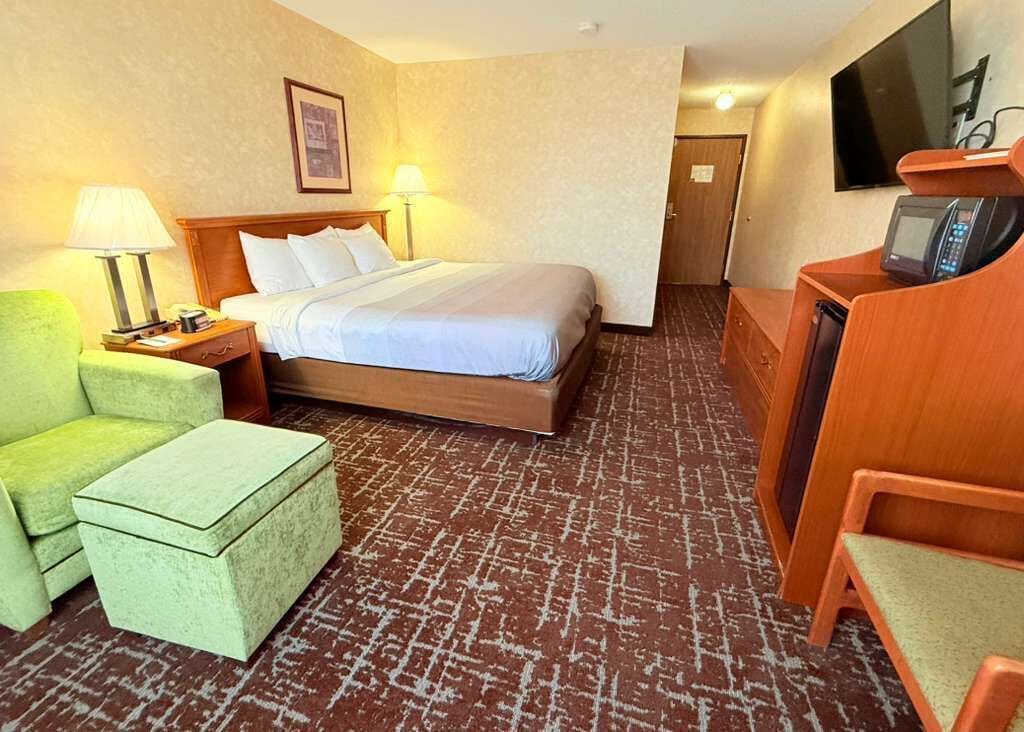 Quarto de hotel com cama, escrivaninha, cadeira, luminária e televisão.