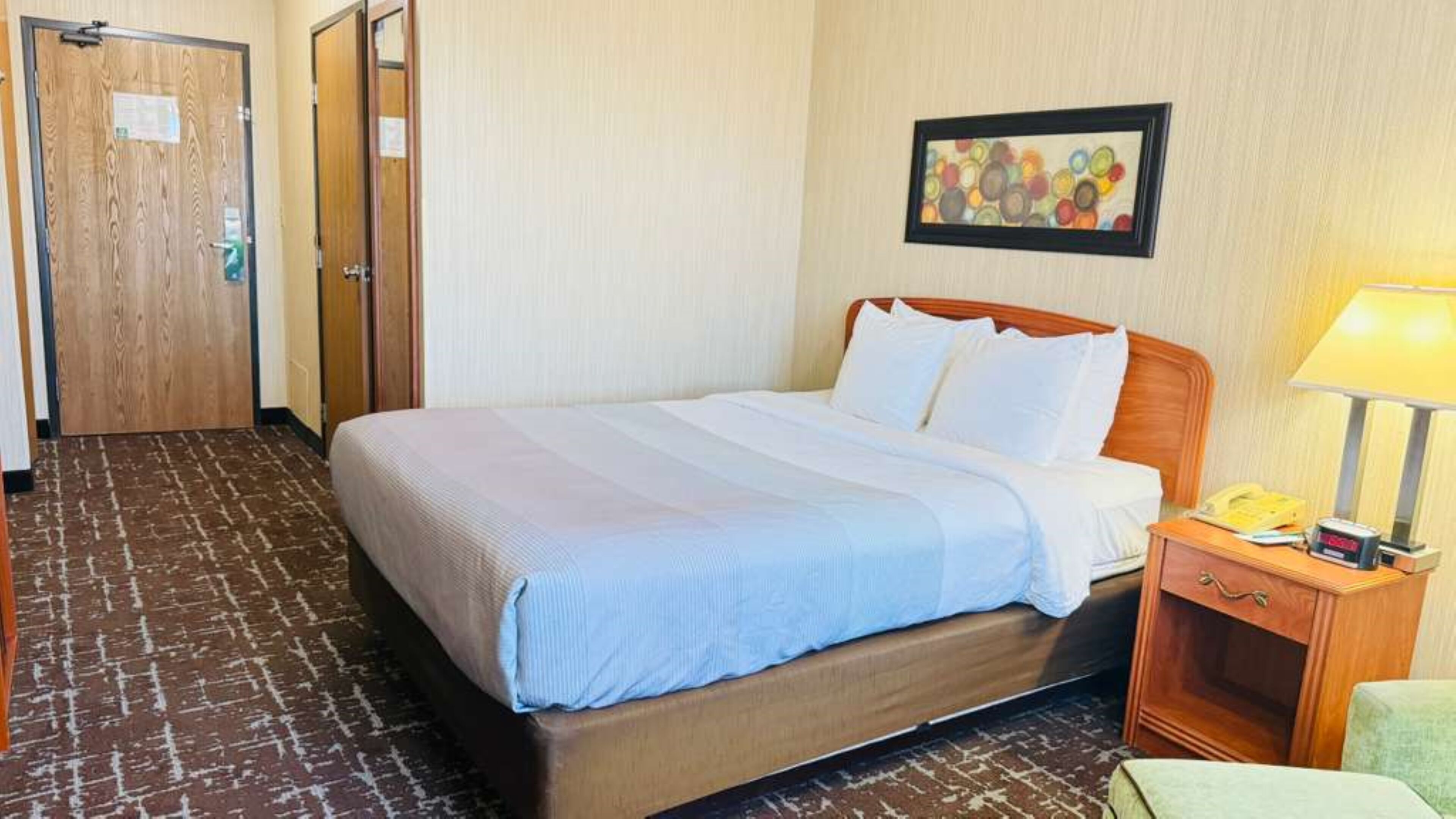 A hotel room with a bed, a bedside table with a lamp, a wall clock, and a door.