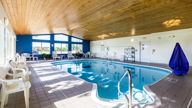An indoor swimming pool with a wooden ceiling, clear view of the outside, and several white chairs around the pool area.
