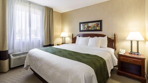 A hotel room with a large bed, two bedside tables with lamps, a window with curtains, a wall-mounted painting, and a thermostat.
