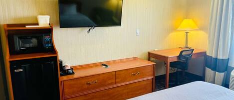 A hotel room with a bed, a desk, a chair, a TV, a microwave, and a lamp.