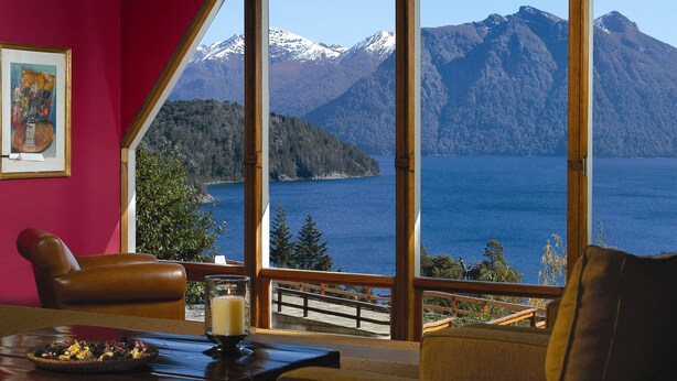 Una habitación con un gran ventanal que da a un lago y montañas. Hay una mesa con una vela y un florero.