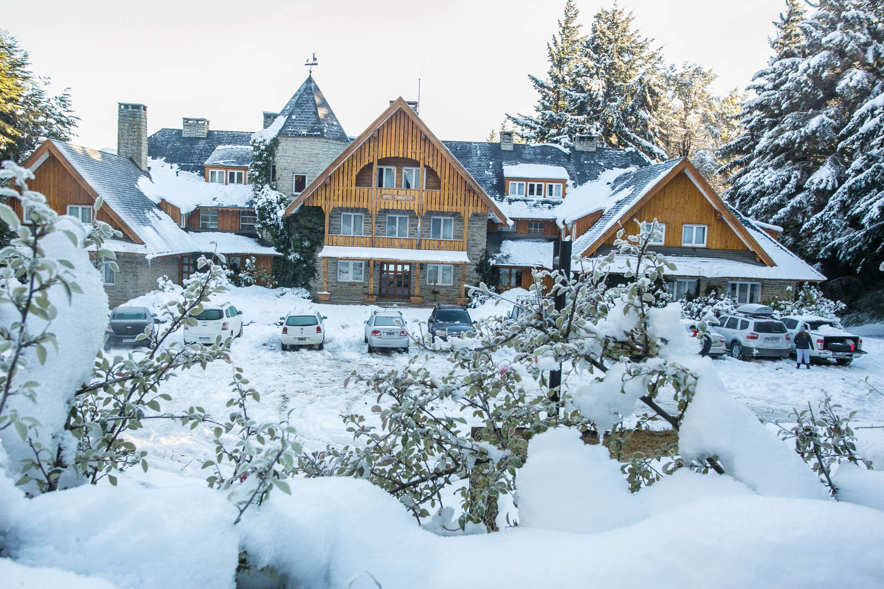 A large wooden lodge with multiple stories and a prominent chimney, surrounded by snow-covered trees and parked cars.