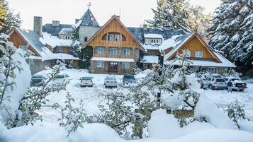 A large wooden lodge with multiple stories and a prominent chimney, surrounded by snow-covered trees and parked cars.