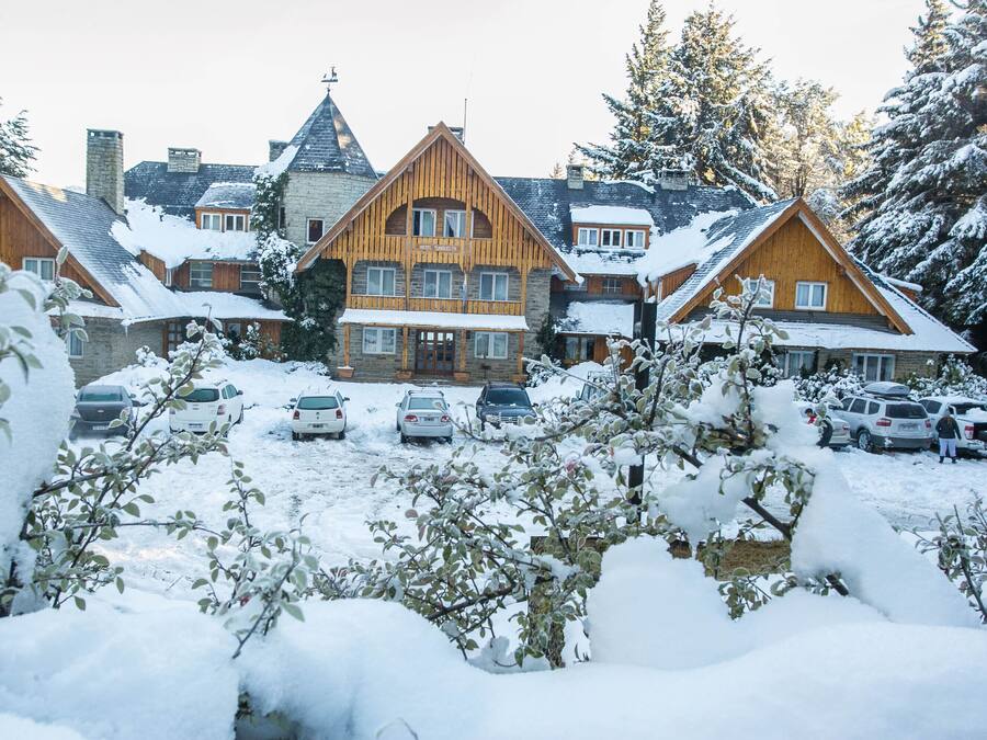 A large wooden lodge with multiple stories and a prominent chimney, surrounded by snow-covered trees and parked cars.