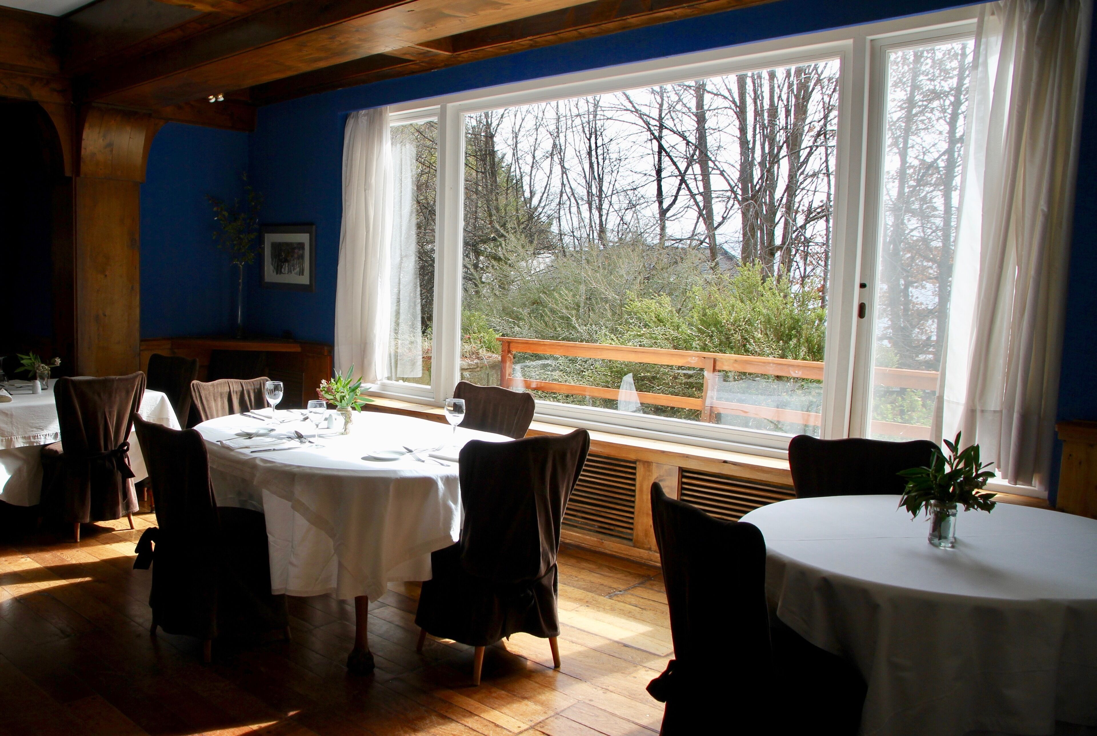 Un espace repas avec des tables rondes, des chaises et une grande fenêtre donnant sur des arbres et une terrasse en bois.