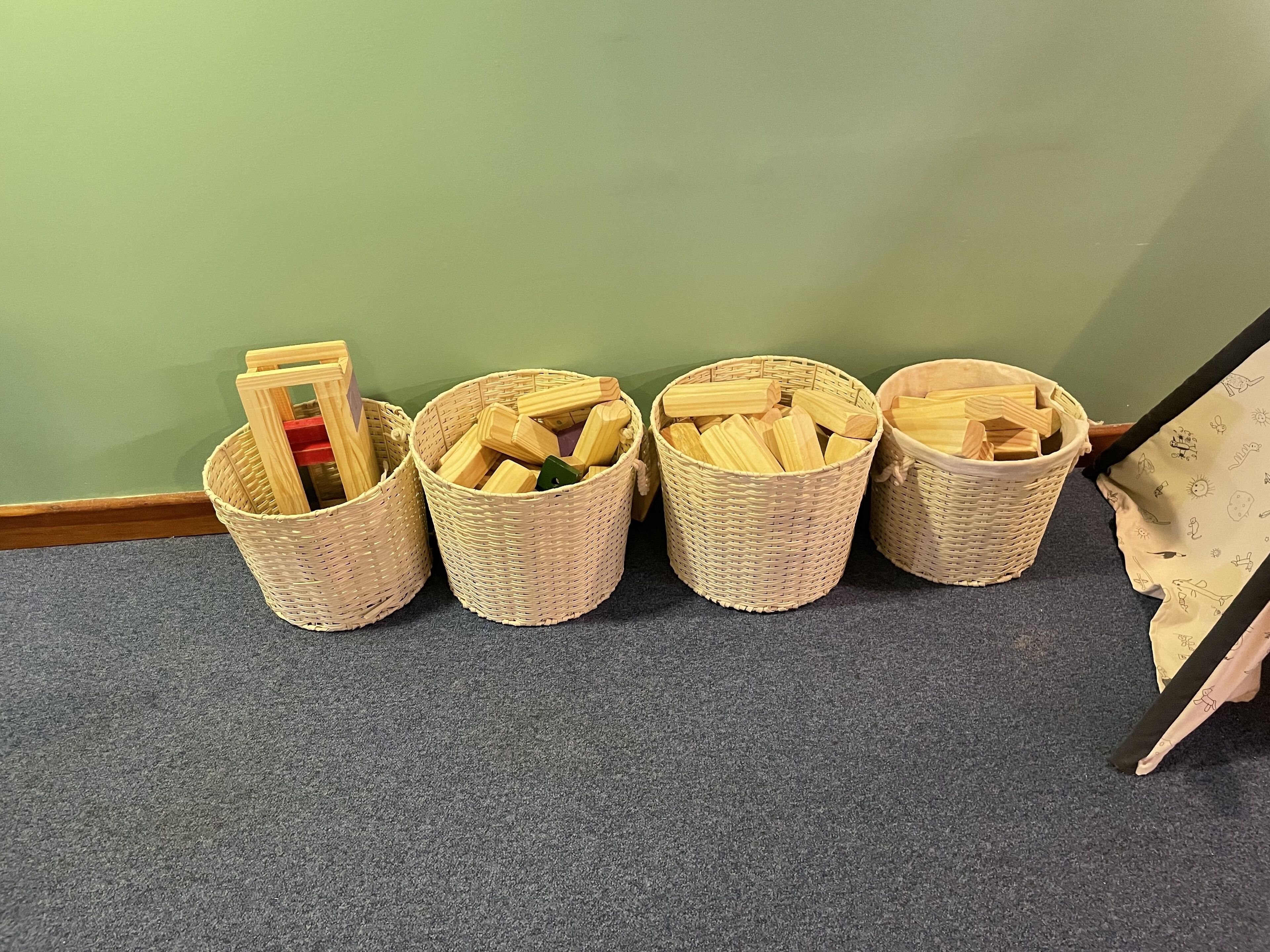 Four wicker baskets filled with wooden blocks on a carpeted floor against a green wall.
