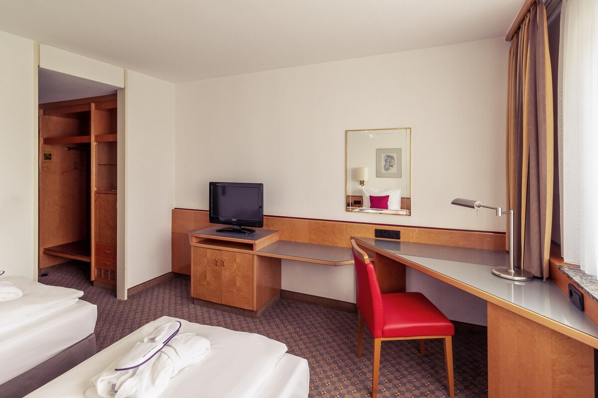 A hotel room with a bed, a desk with a computer, a chair, a wardrobe, and a mirror.