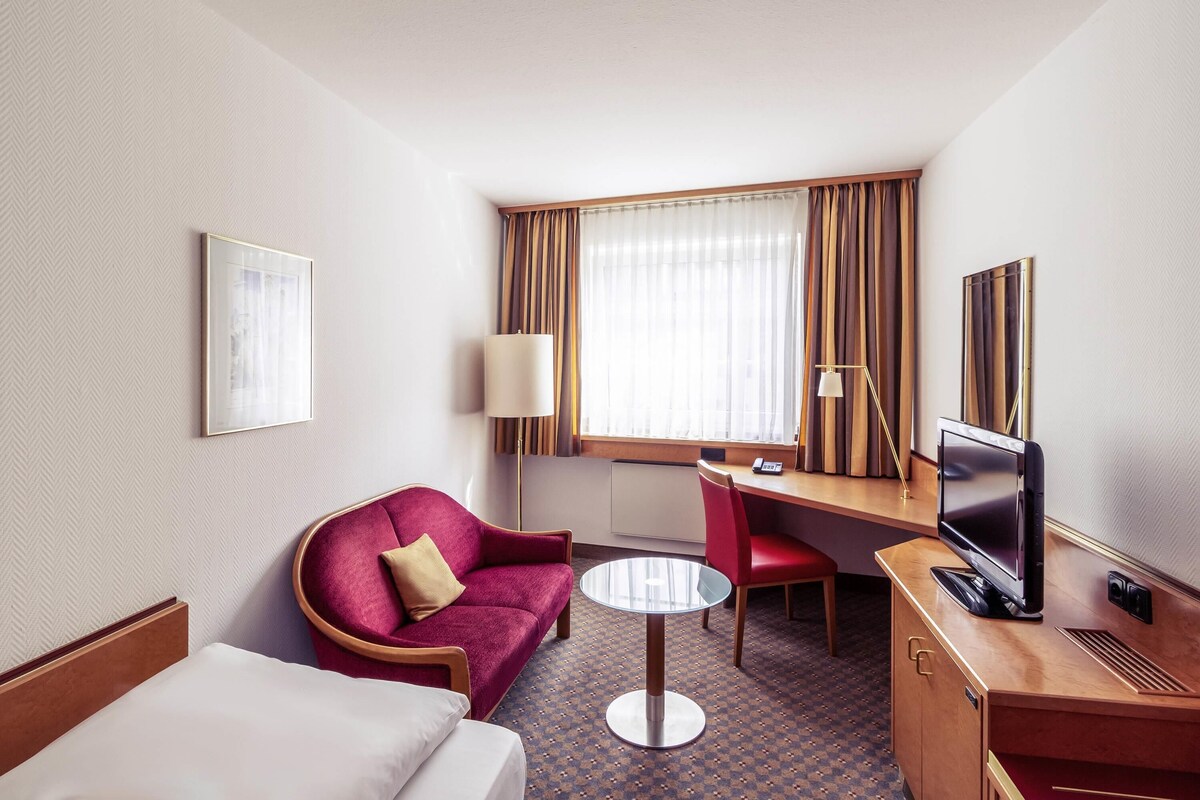 A hotel room with a bed, a red armchair, a desk with a TV, a lamp, and a window with curtains.
