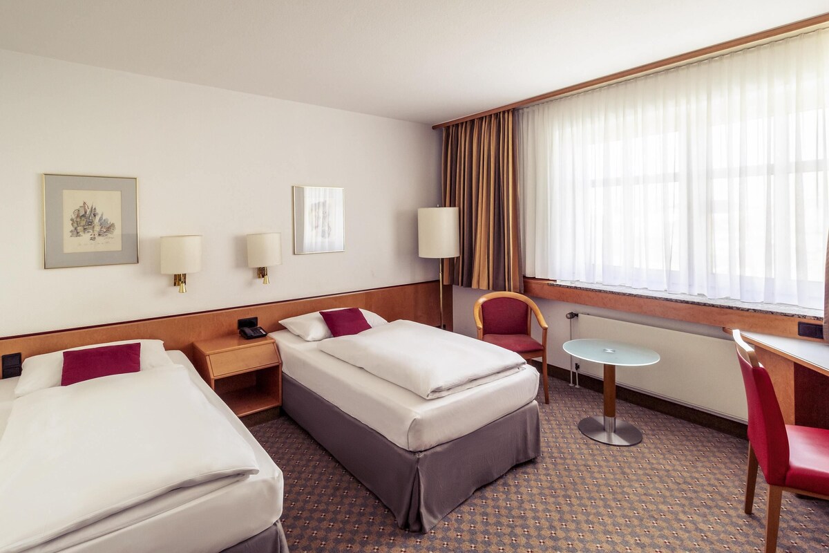 A hotel room with two beds, a desk, a chair, a lamp, and a window with curtains.