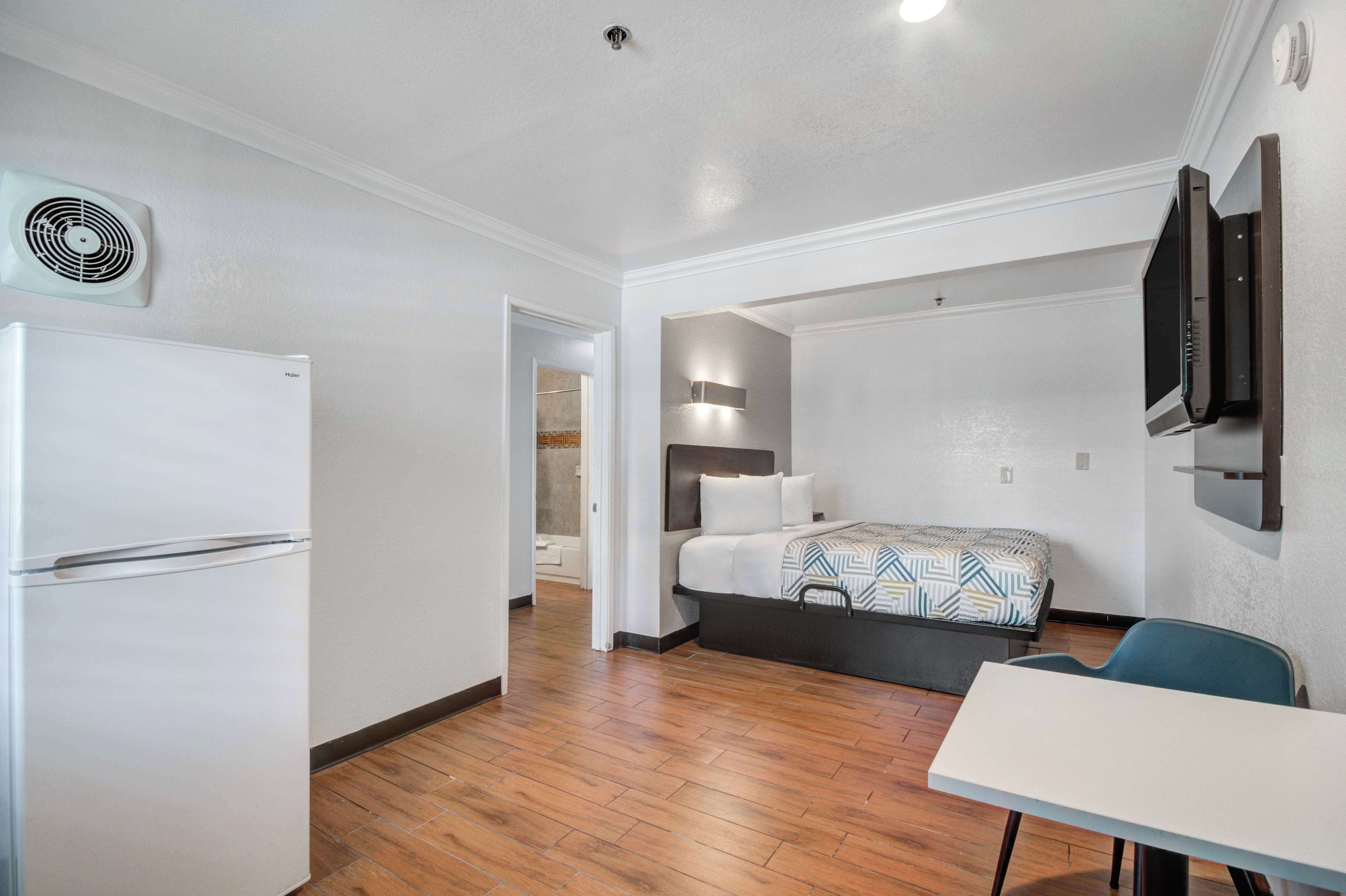 Habitación de hotel compacta con una cama, una mesita, una silla, un televisor montado en la pared y un refrigerador.