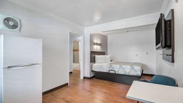Habitación de hotel compacta con una cama, una mesita, una silla, un televisor montado en la pared y un refrigerador.